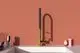 Küchenarmatur von Ideal Standard mit einem gebürsteten Goldfinish. Der Wasserhahn hat einen langen, schlanken Auslauf und befindet sich über einem weißen Waschbecken vor einer rosa Wand.